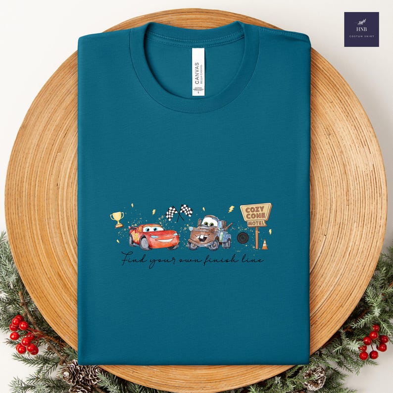 May include: Teal t-shirt featuring a graphic of two cartoon cars, a trophy, and the words "Find your own finish line." The design includes a Cozy Cone Motel sign and checkered flags.
