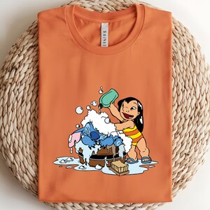 Pode incluir: Camiseta laranja com uma ilustra&ccedil;&atilde;o de desenho animado de Lilo lavando Stitch em uma banheira de madeira. Lilo est&aacute; usando um mai&ocirc; listrado amarelo e laranja. Stitch &eacute; azul. A camisa est&aacute; dobrada em uma superf&iacute;cie tecida.