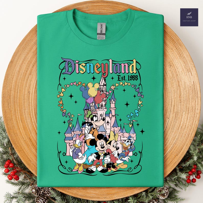 Może przedstawiać: Jasnozielony t-shirt z kr&oacute;tkim rękawem z nadrukiem graficznym Disneyland Est. 1955, przedstawiającym r&oacute;żowy zamek, Myszkę Miki, Myszkę Minnie, Kaczora Donalda, Daisy Duck i Goofy'ego z balonami, w ozdobnej czarnej ramce.