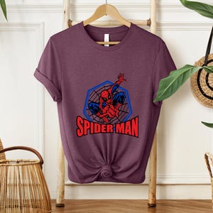 May include: A maroon t-shirt featuring a graphic of Spider-Man in a blue web design with the text "SPIDER-MAN" in red. The superhero is depicted in his classic red and blue suit, striking a dynamic pose.