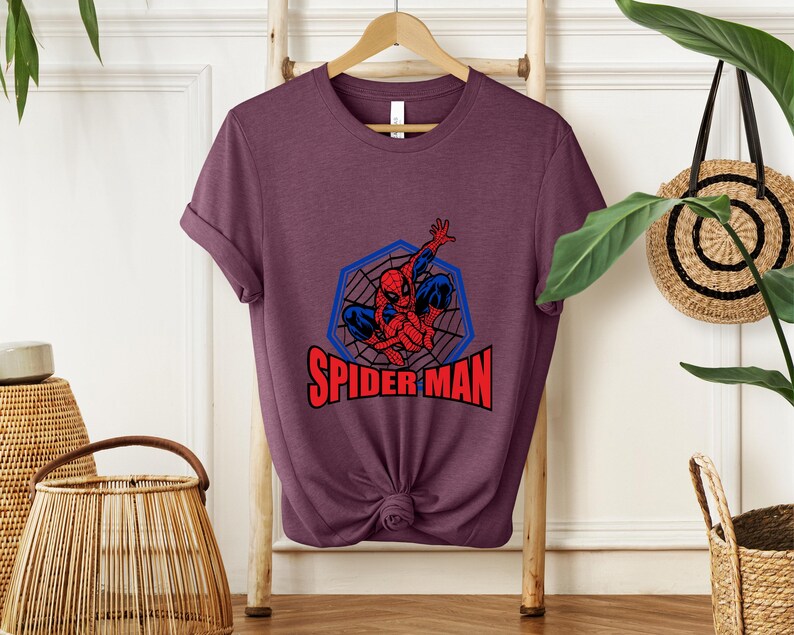 May include: A maroon t-shirt featuring a graphic of Spider-Man in a blue web design with the text "SPIDER-MAN" in red. The superhero is depicted in his classic red and blue suit, striking a dynamic pose.