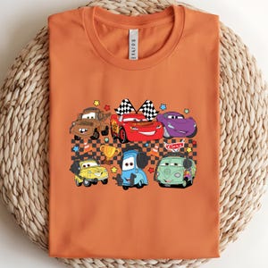 May include: Orange t-shirt featuring a colorful graphic of cartoon cars. The design includes various car characters, checkered flags, and a trophy. The cars are in different colors, including red, blue, yellow, and purple.