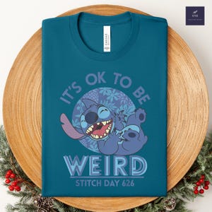Puede incluir: Camiseta de color turquesa con un dise&ntilde;o de Stitch de dibujos animados, el texto dice "IT'S OK TO BE WEIRD" y "STITCH DAY 626". El dise&ntilde;o incluye a Stitch sonriendo y elementos florales. La camiseta se muestra en un plato de madera.