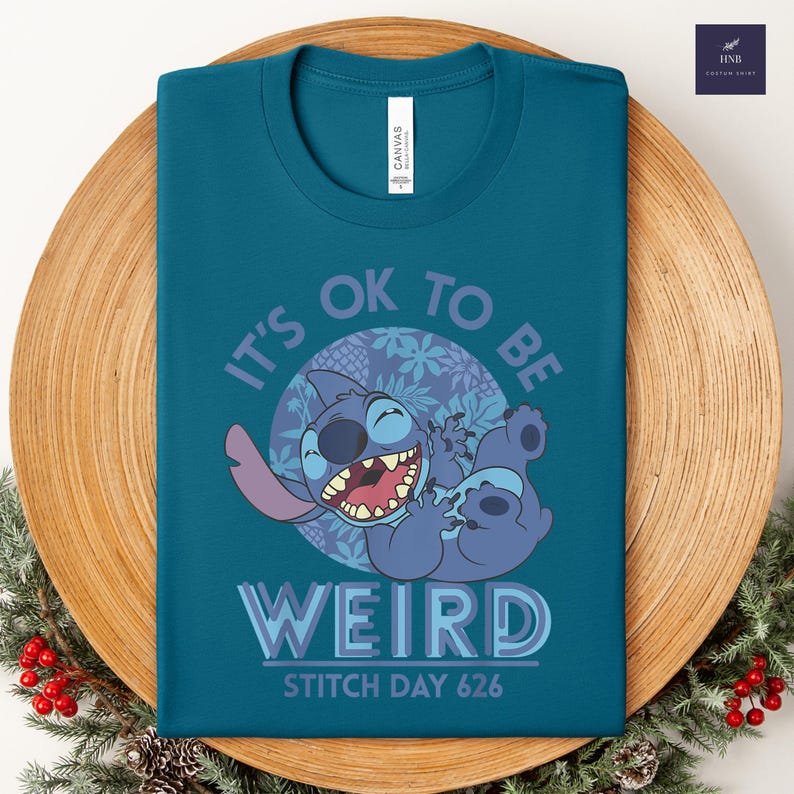 Puede incluir: Camiseta de color turquesa con un dise&ntilde;o de Stitch de dibujos animados, el texto dice "IT'S OK TO BE WEIRD" y "STITCH DAY 626". El dise&ntilde;o incluye a Stitch sonriendo y elementos florales. La camiseta se muestra en un plato de madera.