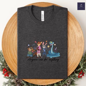 K&ouml;nnte beinhalten: Dunkelgraues T-Shirt mit einer farbenfrohen Cartoon-Illustration von Tierfiguren. Das Design umfasst einen Biber, einen Fuchs, ein Kaninchen und eine Schlange, mit dem Text "Anyone can be anything". Das Shirt ist auf einem Holztablett gefaltet.
