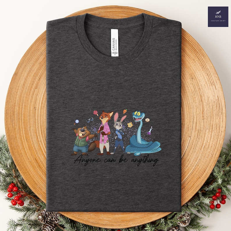 K&ouml;nnte beinhalten: Dunkelgraues T-Shirt mit einer farbenfrohen Cartoon-Illustration von Tierfiguren. Das Design umfasst einen Biber, einen Fuchs, ein Kaninchen und eine Schlange, mit dem Text "Anyone can be anything". Das Shirt ist auf einem Holztablett gefaltet.