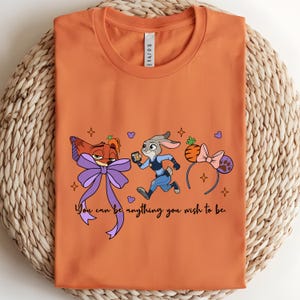 May include: Orange t-shirt featuring cartoon characters and the text "You can be anything you wish to be." The design includes a fox with a purple bow, a rabbit in a police uniform, and a headband with mouse ears.