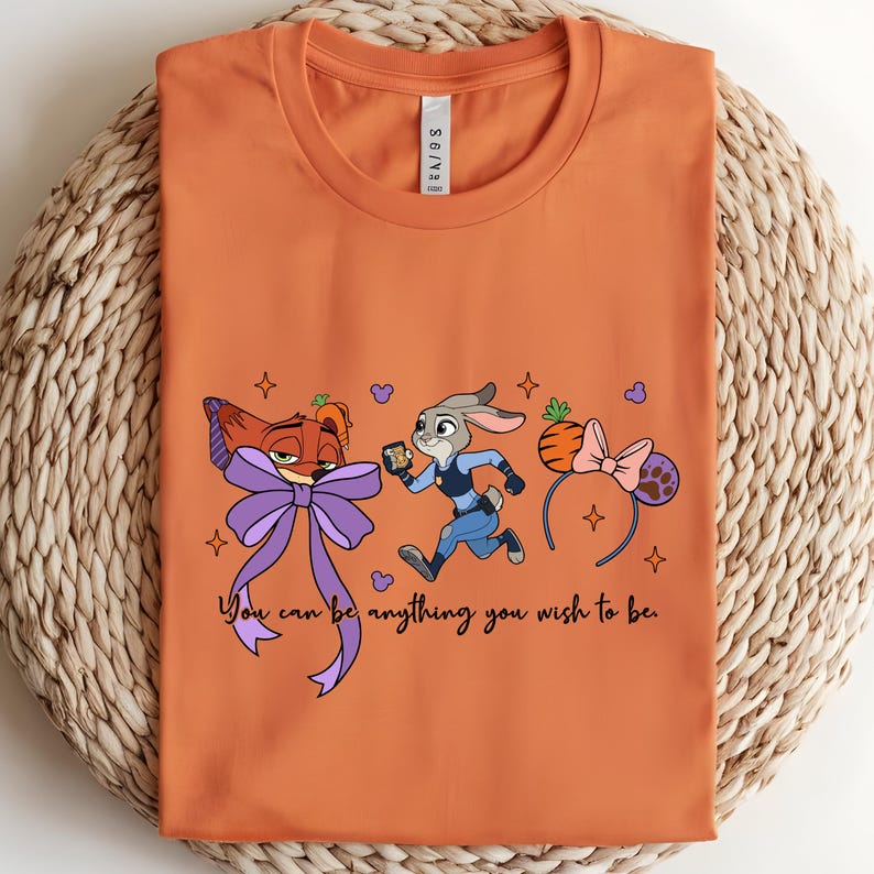 May include: Orange t-shirt featuring cartoon characters and the text "You can be anything you wish to be." The design includes a fox with a purple bow, a rabbit in a police uniform, and a headband with mouse ears.