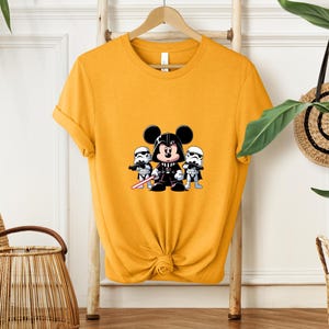 Pu&ograve; includere: T-shirt giallo senape con una grafica a cartoni animati di Topolino vestito da Darth Vader, affiancato da due Stormtrooper. La grafica &egrave; in nero, bianco e rosso. La maglietta &egrave; appesa a un supporto di legno.