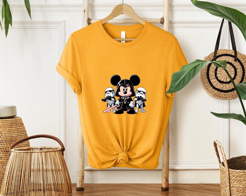 Pu&ograve; includere: T-shirt giallo senape con una grafica a cartoni animati di Topolino vestito da Darth Vader, affiancato da due Stormtrooper. La grafica &egrave; in nero, bianco e rosso. La maglietta &egrave; appesa a un supporto di legno.