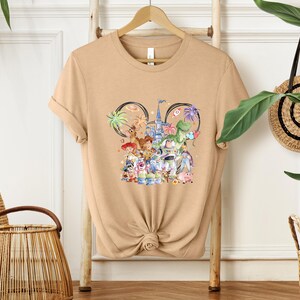 May include: Tan t-shirt featuring a colorful graphic of Disney characters, including Woody, Buzz Lightyear, and others, with a castle and Mickey Mouse ears design. The shirt is displayed on a wooden hanger.