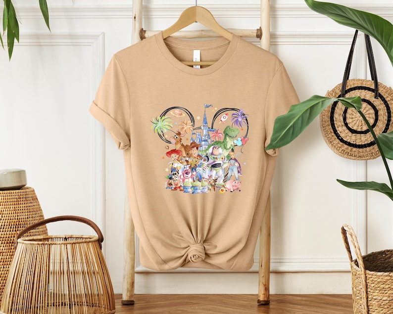 May include: Tan t-shirt featuring a colorful graphic of Disney characters, including Woody, Buzz Lightyear, and others, with a castle and Mickey Mouse ears design. The shirt is displayed on a wooden hanger.