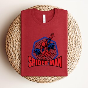 May include: A red t-shirt featuring a graphic of Spider-Man in a blue and red design, with the words "SPIDER MAN" in red block letters. The shirt is folded and placed on a woven, circular mat.