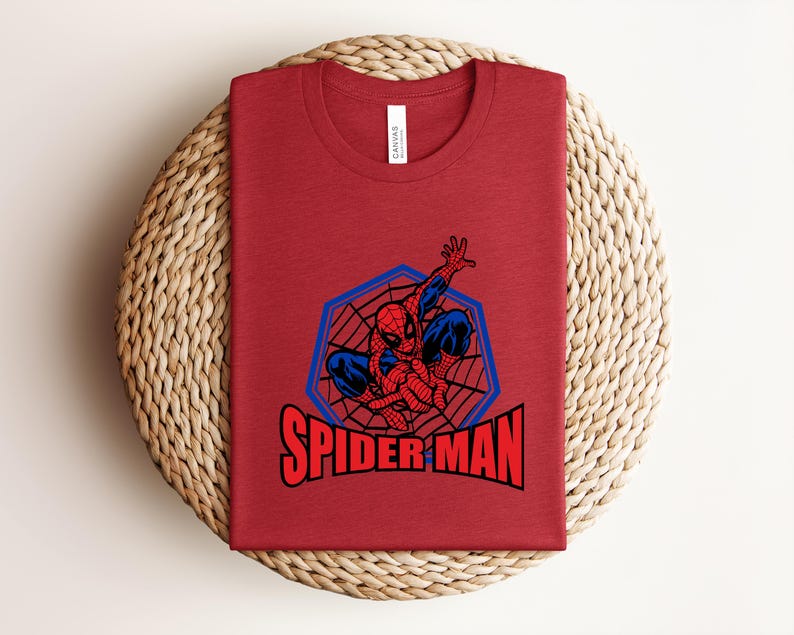 May include: A red t-shirt featuring a graphic of Spider-Man in a blue and red design, with the words "SPIDER MAN" in red block letters. The shirt is folded and placed on a woven, circular mat.