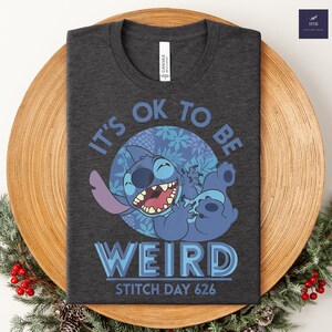 Puede incluir: Camiseta gris oscuro con un personaje de dibujos animados azul y el texto "IT'S OK TO BE WEIRD" y "STITCH DAY 626". El dise&ntilde;o utiliza tonos azules y morados.