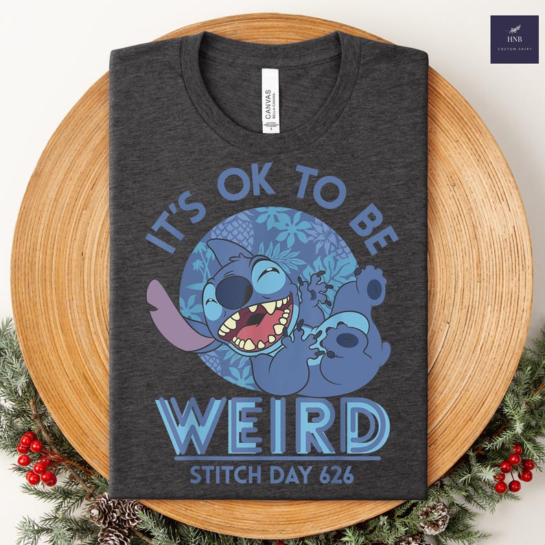Puede incluir: Camiseta gris oscuro con un personaje de dibujos animados azul y el texto "IT'S OK TO BE WEIRD" y "STITCH DAY 626". El dise&ntilde;o utiliza tonos azules y morados.