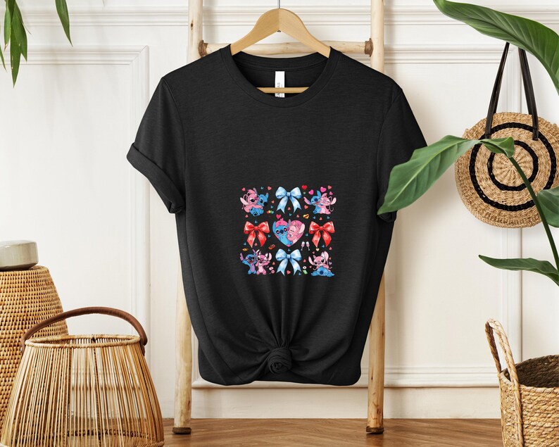 May include: Black t-shirt featuring a colorful graphic of cartoon characters, bows, and hearts. The design includes blue, pink, and red elements. The shirt is hanging on a wooden rack.
