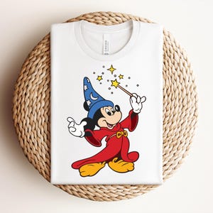 Puede incluir: Camiseta blanca con una ilustraci&oacute;n de dibujos animados de Mickey Mouse vestido de mago. Mickey lleva un sombrero azul con estrellas, una t&uacute;nica roja y zapatos amarillos, sosteniendo una varita con estrellas. La camiseta est&aacute; doblada sobre una superficie circular tejida.