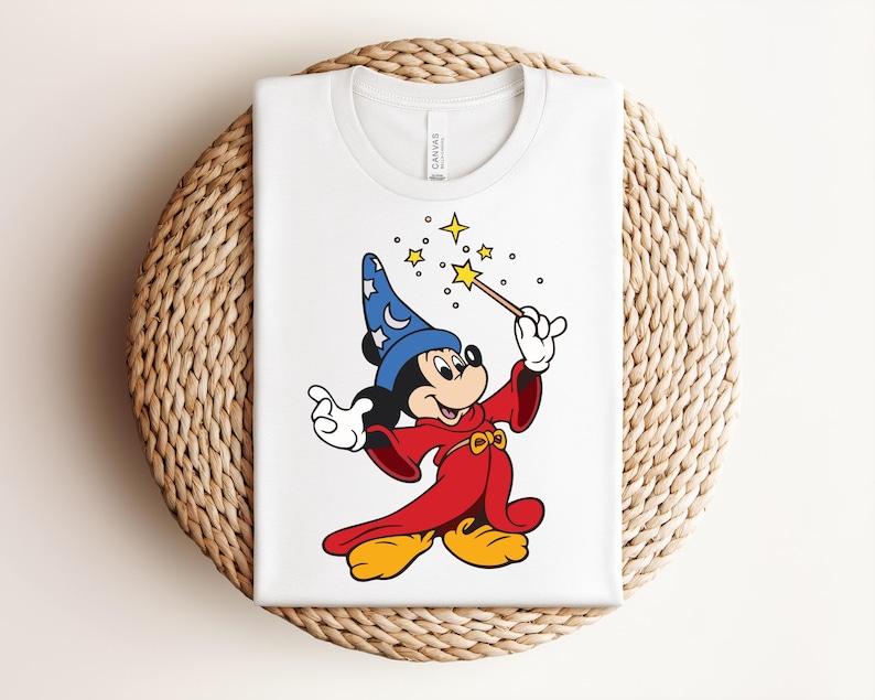 Puede incluir: Camiseta blanca con una ilustraci&oacute;n de dibujos animados de Mickey Mouse vestido de mago. Mickey lleva un sombrero azul con estrellas, una t&uacute;nica roja y zapatos amarillos, sosteniendo una varita con estrellas. La camiseta est&aacute; doblada sobre una superficie circular tejida.