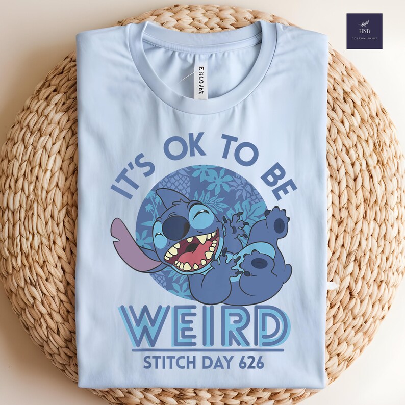 Puede incluir: Camiseta azul claro con una ilustraci&oacute;n de Stitch. El dise&ntilde;o incluye el texto "IT'S OK TO BE WEIRD" y "STITCH DAY 626". La imagen es una ilustraci&oacute;n juguetona del personaje.
