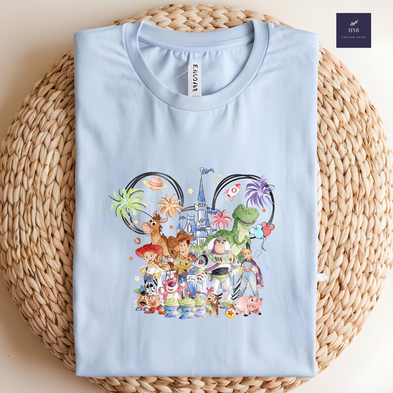 May include: Light blue t-shirt featuring a colorful graphic of Disney's Toy Story characters within a Mickey Mouse ear outline. The design includes Woody, Buzz Lightyear, and other characters. The shirt is folded on a woven surface.