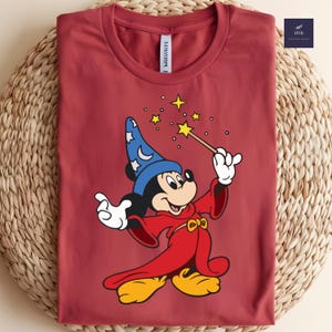 Puede incluir: Una camiseta granate con Mickey Mouse de dibujos animados vestido de mago, sosteniendo una varita con una estrella al final. Mickey lleva un sombrero de mago azul y t&uacute;nicas rojas. La camiseta est&aacute; doblada sobre una superficie tejida.