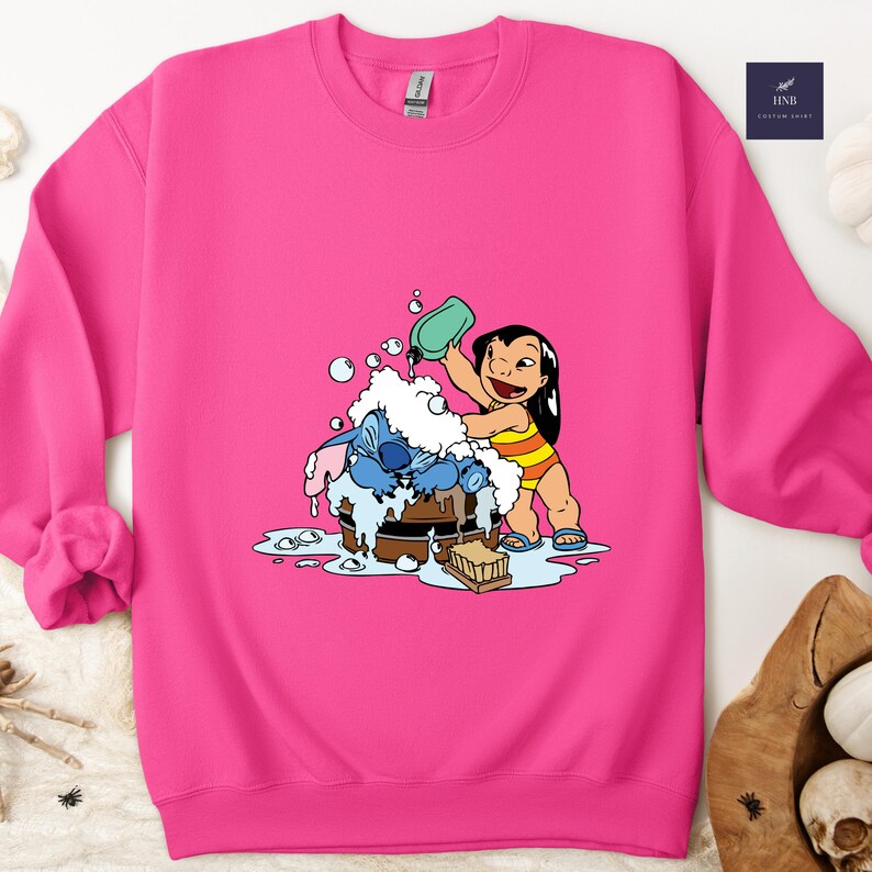 Pode incluir: Um moletom rosa brilhante com uma ilustra&ccedil;&atilde;o de desenho animado de Lilo e Stitch. Lilo est&aacute; derramando &aacute;gua sobre Stitch, que est&aacute; em um barril de madeira cheio de bolhas. O design &eacute; em estilo cartoon.