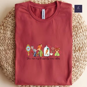 May include: A maroon t-shirt featuring a cartoon design with the text "You are my happily ever after." The design includes a mirror, a bunny, a fox, a carrot in a glass dome, roses, and a teacup. The shirt is made of soft material.