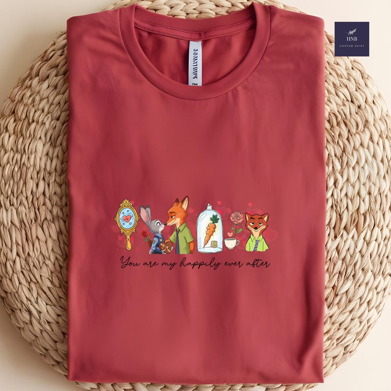 May include: A maroon t-shirt featuring a cartoon design with the text "You are my happily ever after." The design includes a mirror, a bunny, a fox, a carrot in a glass dome, roses, and a teacup. The shirt is made of soft material.