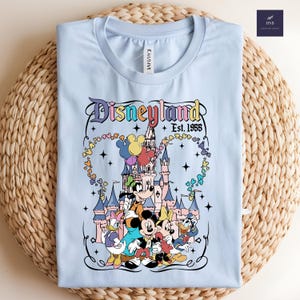 Może przedstawiać: Jasnoniebieski t-shirt z grafiką przedstawiającą zamek, Myszkę Miki, Myszkę Minnie, Goofy'ego, Kaczora Donalda i Daisy. Tekst głosi Disneyland Est. 1955. Wz&oacute;r zawiera kolorowe balony, gwiazdki oraz ozdobną ramkę wok&oacute;ł ilustracji.