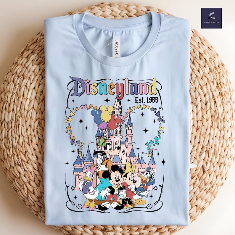 Może przedstawiać: Jasnoniebieski t-shirt z grafiką przedstawiającą zamek, Myszkę Miki, Myszkę Minnie, Goofy'ego, Kaczora Donalda i Daisy. Tekst głosi Disneyland Est. 1955. Wz&oacute;r zawiera kolorowe balony, gwiazdki oraz ozdobną ramkę wok&oacute;ł ilustracji.
