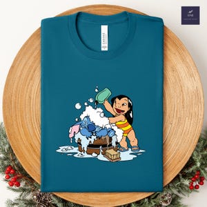 Pode incluir: Camiseta azul-petr&oacute;leo com uma ilustra&ccedil;&atilde;o de desenho animado de Lilo lavando Stitch em uma banheira de madeira. Lilo est&aacute; usando um mai&ocirc; listrado amarelo e laranja. A camisa tem a palavra "CANVAS" impressa.