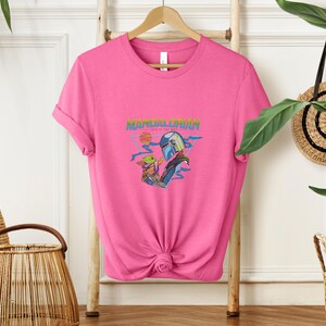 May include: A vibrant pink t-shirt with a graphic of The Mandalorian and Grogu. The design includes the text "MANDALORIAN" in a retro font, with a sunset and space-themed elements. The shirt is knotted at the bottom.
