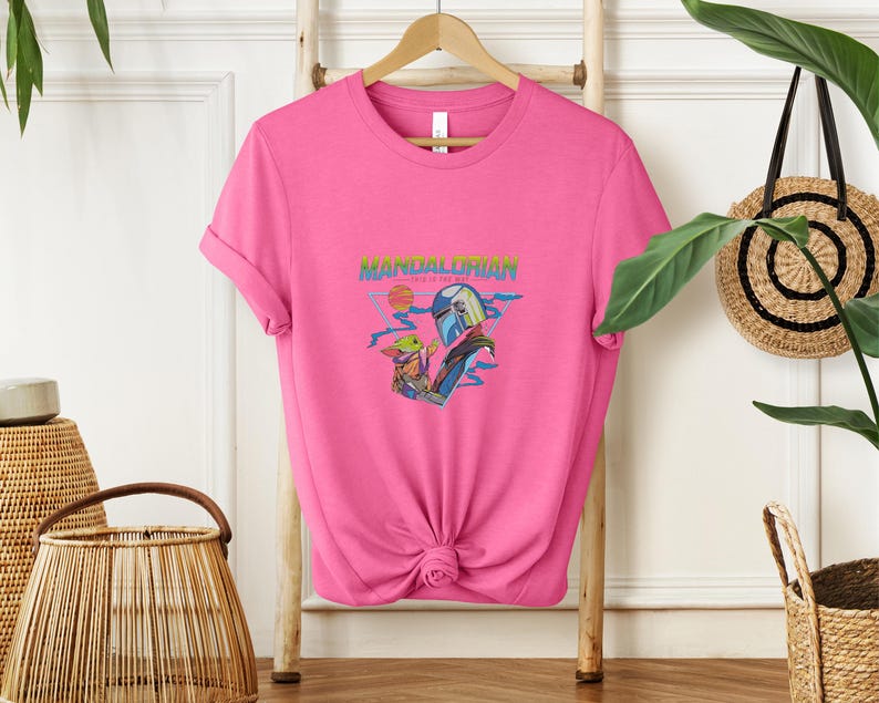 May include: A vibrant pink t-shirt with a graphic of The Mandalorian and Grogu. The design includes the text "MANDALORIAN" in a retro font, with a sunset and space-themed elements. The shirt is knotted at the bottom.