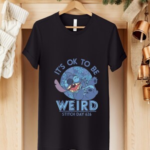 Puede incluir: Camiseta negra con un dise&ntilde;o de Stitch de Lilo & Stitch. El dise&ntilde;o muestra a Stitch en una pose juguetona, con el texto "IT'S OK TO BE WEIRD" y "STITCH DAY 626". El fondo es un patr&oacute;n floral azul.
