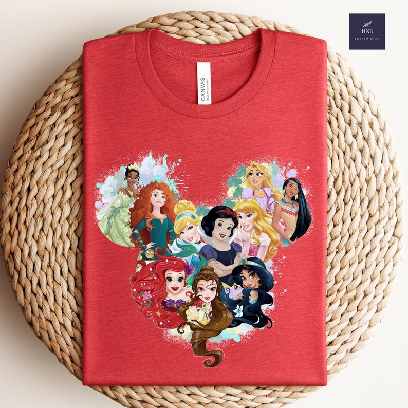 K&ouml;nnte beinhalten: Ein rotes T-Shirt mit einer Grafik von Disney-Prinzessinnen, die in Form eines Mickey-Mouse-Kopfes angeordnet sind. Das Design umfasst Arielle, Belle, Schneewittchen und andere Prinzessinnen. Das T-Shirt hat ein "Canvas"-Etikett.