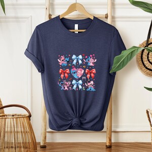 May include: Navy blue t-shirt featuring a colorful graphic of cartoon characters, hearts, and bows. The design includes characters in various poses, with red, pink, and blue accents. The shirt is made of a soft material and has a classic crew neck.
