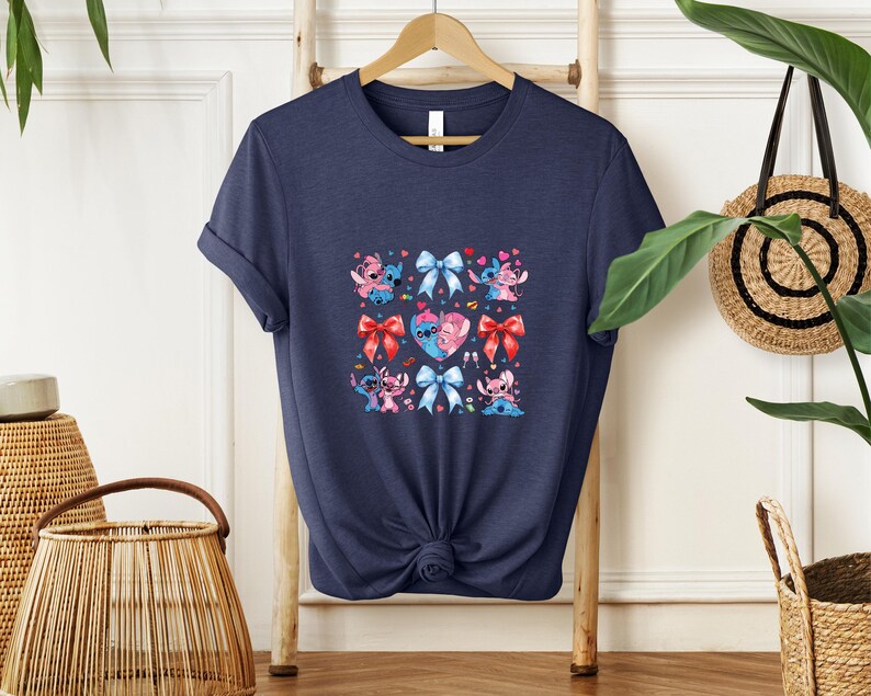 May include: Navy blue t-shirt featuring a colorful graphic of cartoon characters, hearts, and bows. The design includes characters in various poses, with red, pink, and blue accents. The shirt is made of a soft material and has a classic crew neck.