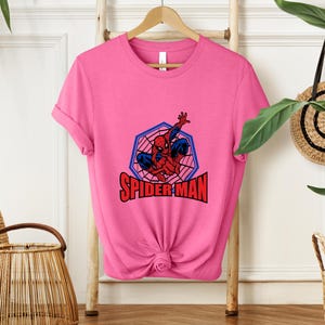 May include: A pink t-shirt featuring a Spider-Man graphic. The design includes a red and blue Spider-Man figure within a blue and red web-shaped frame, with the words "SPIDER-MAN" in red block letters.