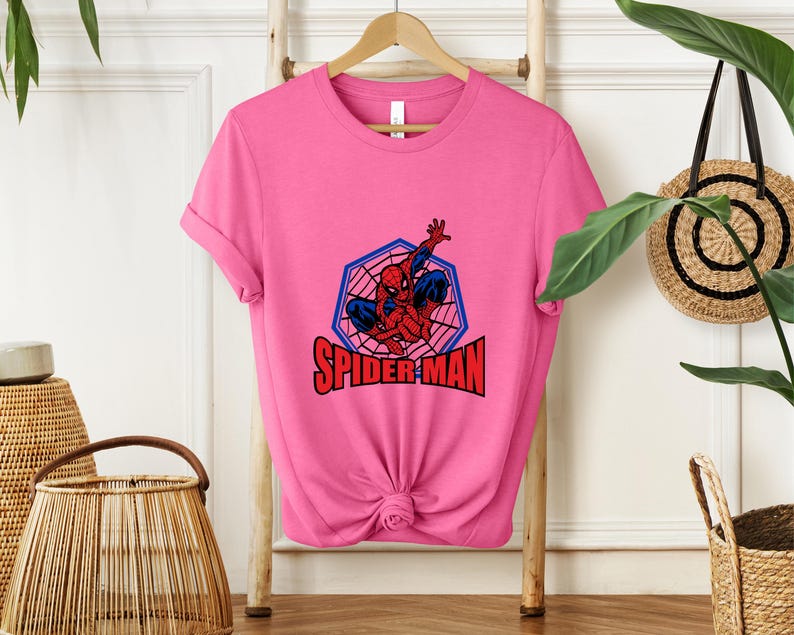 May include: A pink t-shirt featuring a Spider-Man graphic. The design includes a red and blue Spider-Man figure within a blue and red web-shaped frame, with the words "SPIDER-MAN" in red block letters.