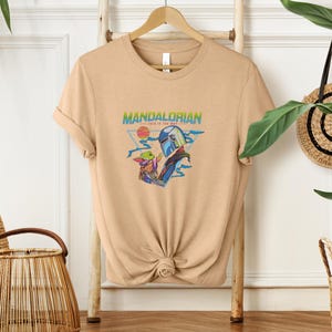 May include: A tan t-shirt with a graphic of The Mandalorian and Grogu. The design includes the text "MANDALORIAN" and "THIS IS THE WAY." The artwork features the characters with a sunset and space-themed elements.