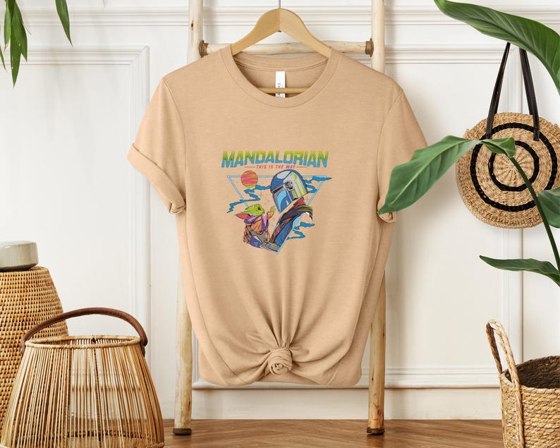 May include: A tan t-shirt with a graphic of The Mandalorian and Grogu. The design includes the text "MANDALORIAN" and "THIS IS THE WAY." The artwork features the characters with a sunset and space-themed elements.