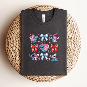 May include: Dark gray t-shirt featuring a colorful graphic of cartoon characters, bows, and hearts. The design includes blue, pink, and red elements. The shirt is displayed on a woven, circular surface.