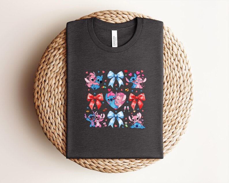 May include: Dark gray t-shirt featuring a colorful graphic of cartoon characters, bows, and hearts. The design includes blue, pink, and red elements. The shirt is displayed on a woven, circular surface.