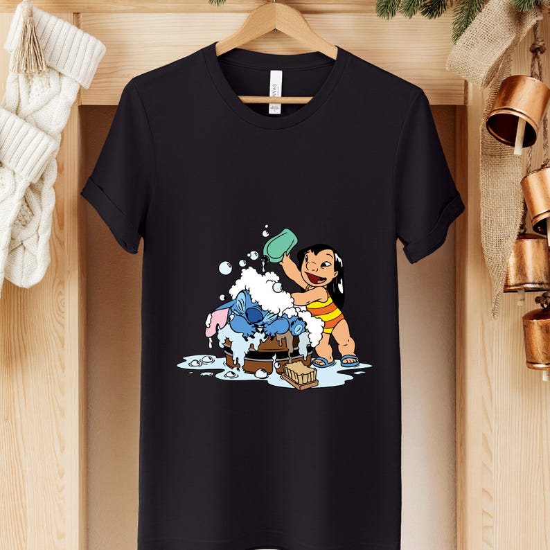 Pode incluir: Camiseta preta com uma ilustra&ccedil;&atilde;o de desenho animado de Lilo lavando Stitch. Lilo est&aacute; usando um mai&ocirc; listrado amarelo e laranja. Stitch &eacute; azul e est&aacute; sendo lavado em uma tina de madeira com bolhas. O desenho est&aacute; centralizado na camiseta.