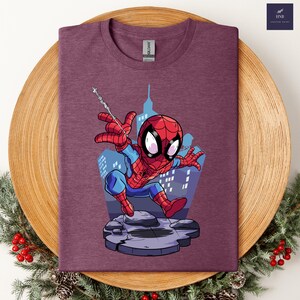 May include: Maroon t-shirt featuring a cartoon Spider-Man graphic. The superhero is depicted in his classic red and blue suit, poised in a dynamic pose with a web. The background shows a cityscape. The shirt has a crew neck and a Gildan tag.