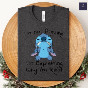 May include: Dark gray t-shirt featuring a blue cartoon character with the text "I'm not Arguing I'm Explaining why I'm Right". The shirt is made of a soft material.