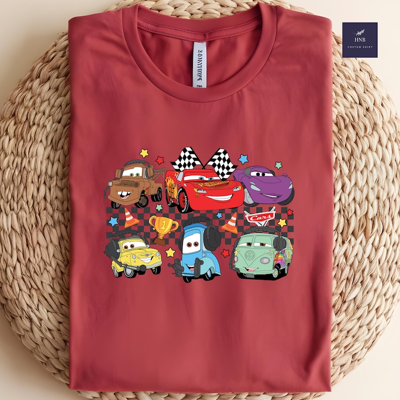 May include: A maroon t-shirt featuring a colorful graphic of cartoon cars, including a red race car with a checkered flag, a yellow car, and a blue car. The design includes stars, a trophy, and the word "Cars".