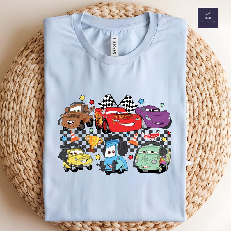 May include: A light blue t-shirt featuring a colorful graphic of cartoon cars. The design includes various car characters, a checkered flag, and a trophy. The word "Cars" is also visible in the design.
