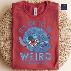 Puede incluir: Camiseta roja con un personaje de dibujos animados azul y el texto "IT'S OK TO BE WEIRD" y "STITCH DAY 626". El personaje est&aacute; sonriendo y tumbado. La camiseta est&aacute; doblada sobre una superficie tejida.